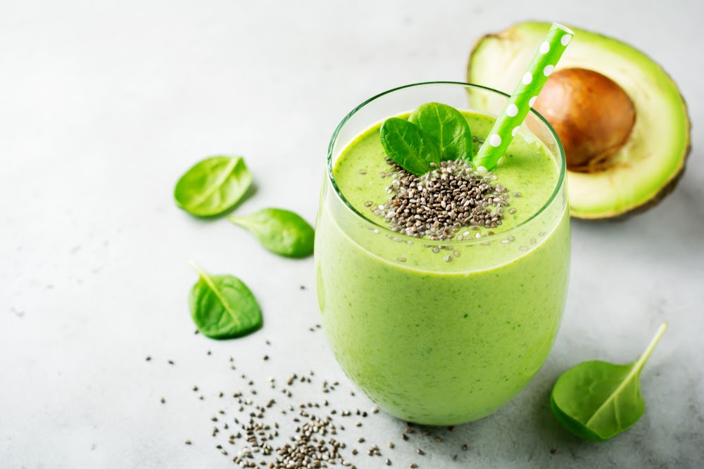 A close up shot of a green smoothie in a glass topped with a basil and chia seed garnish. A white backdrop with some basil leaves, chia seed, and an avocado.