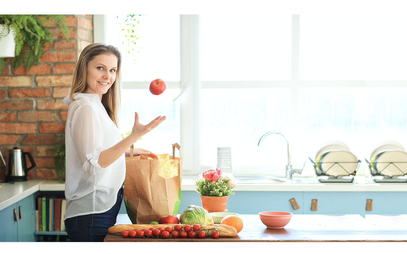 Image of a woman standing in front of a wooden table on her kitchen joggling an apple on her right hand. On the wooden table top is her groceries.