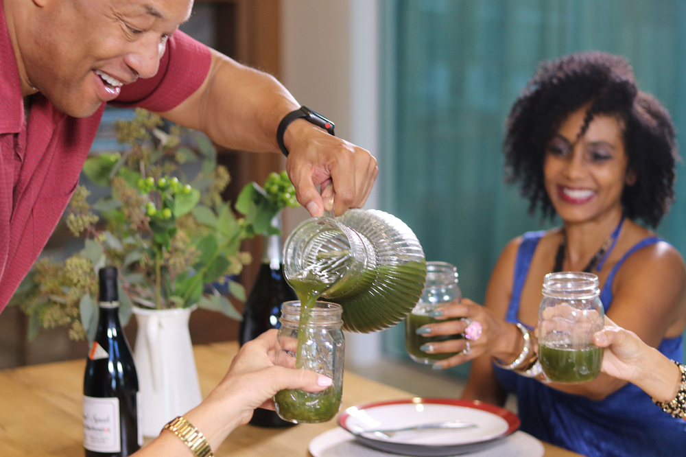 A middle-aged man carrying a pitcher of green smoothie and pouring some to his friends' glass. Set in a dining table.
