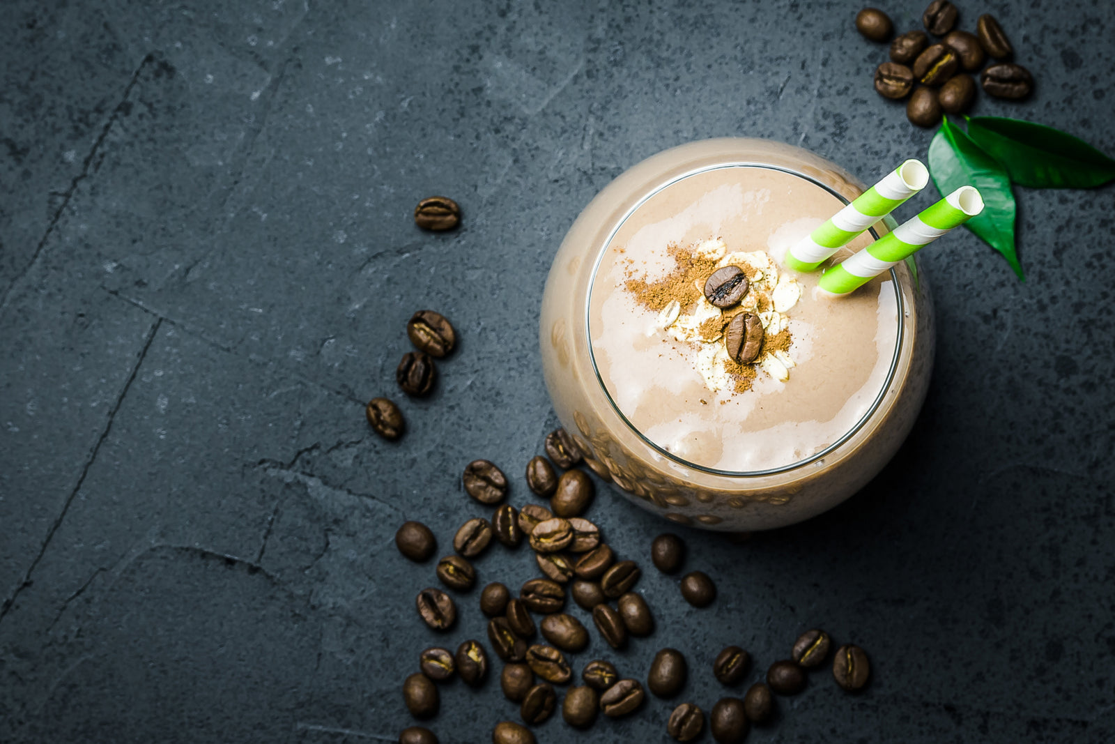 top view shot of a glass of Proteini Iced Coffee with 2 straws topped with 2 coffee beans and on the surface is more coffee beans.