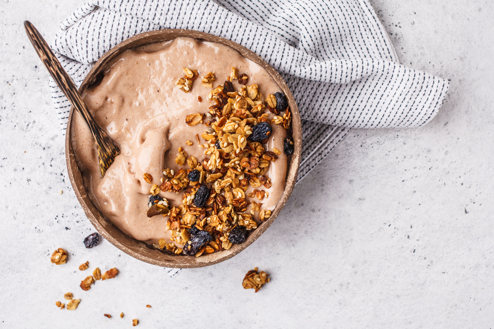 top view shot of a bowl of Peanut Butter Chocolate Proteini shake topped with granola oats and dried berries with a spoon, on the surface is a light blue cloth.