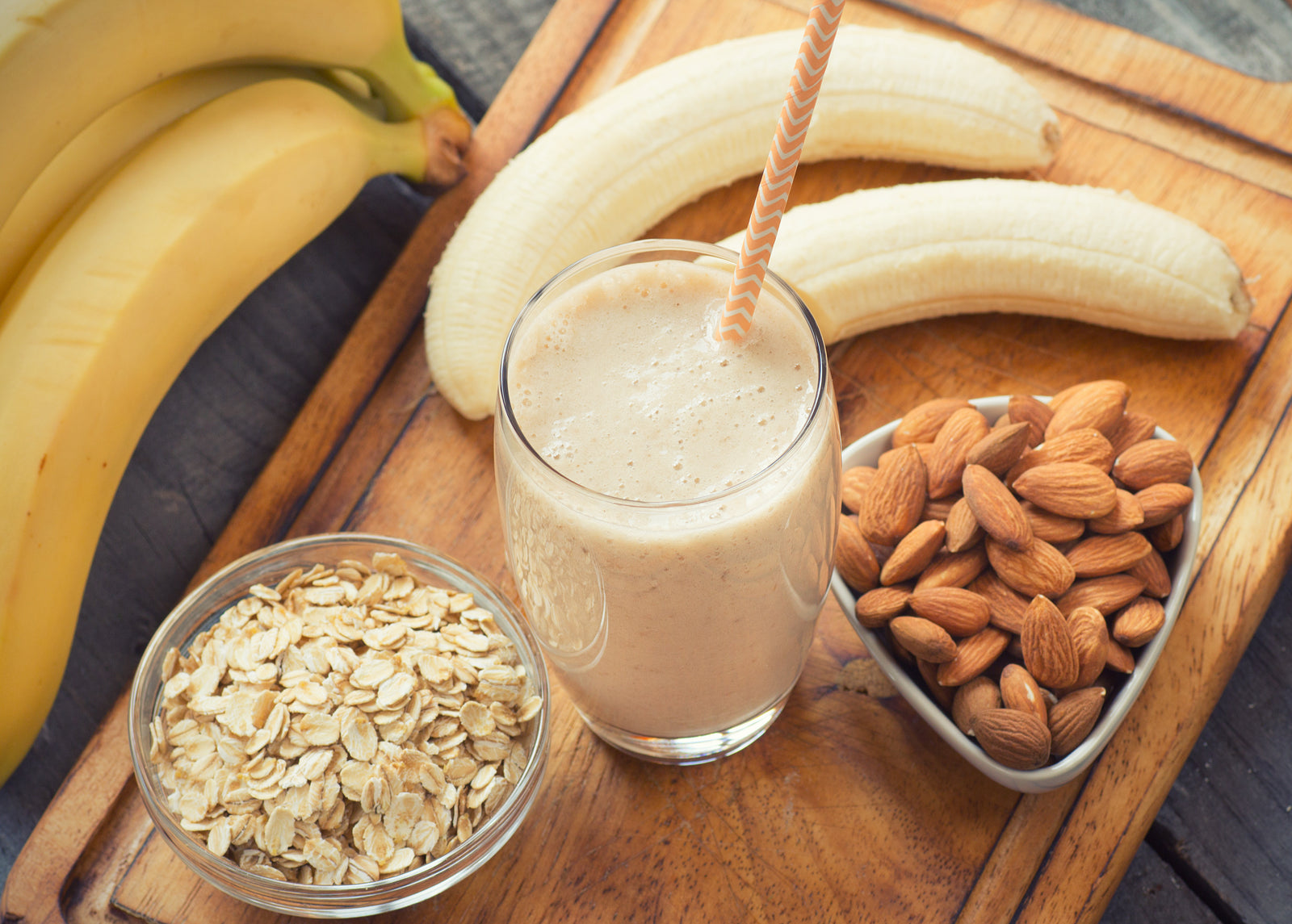 top view shot of a glass of Vegan Vanilla Proteini smoothie with a straw beside 2 peeled bananas, a bowl of almond nuts, a bowl of oats on a wooden cutting boards and beside 3 bananas.