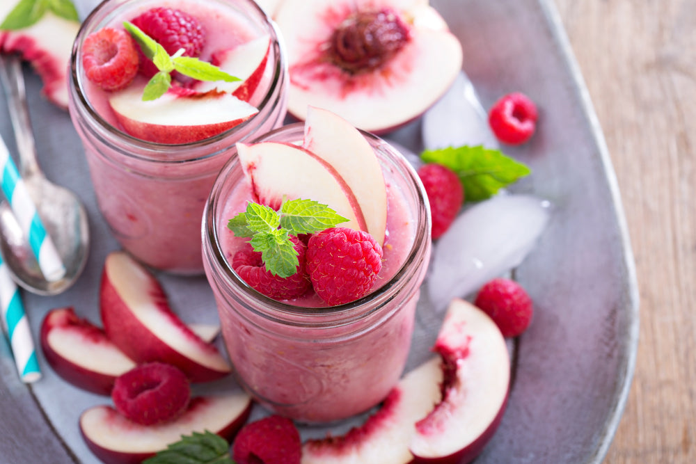 two glasses of smoothie topped with two frozen berries and two slices of peaches garnished with mint leaves surrounded by slices of peaches, berries, and a spoon on a plate