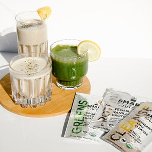1 tall glass of white smoothie with chia seed, a straw, and a slice of pineapple, small glass of white smoothie with green garnish, and a small glass of green smoothie with a slice of lemon. The 3 glasses are all placed in one wooden coaster. Beside is 3 single serving sachets of Organic Pressed Greens, Vegan Vanilla Proteini, and Pineapple Chia Cleanse on a white surface.