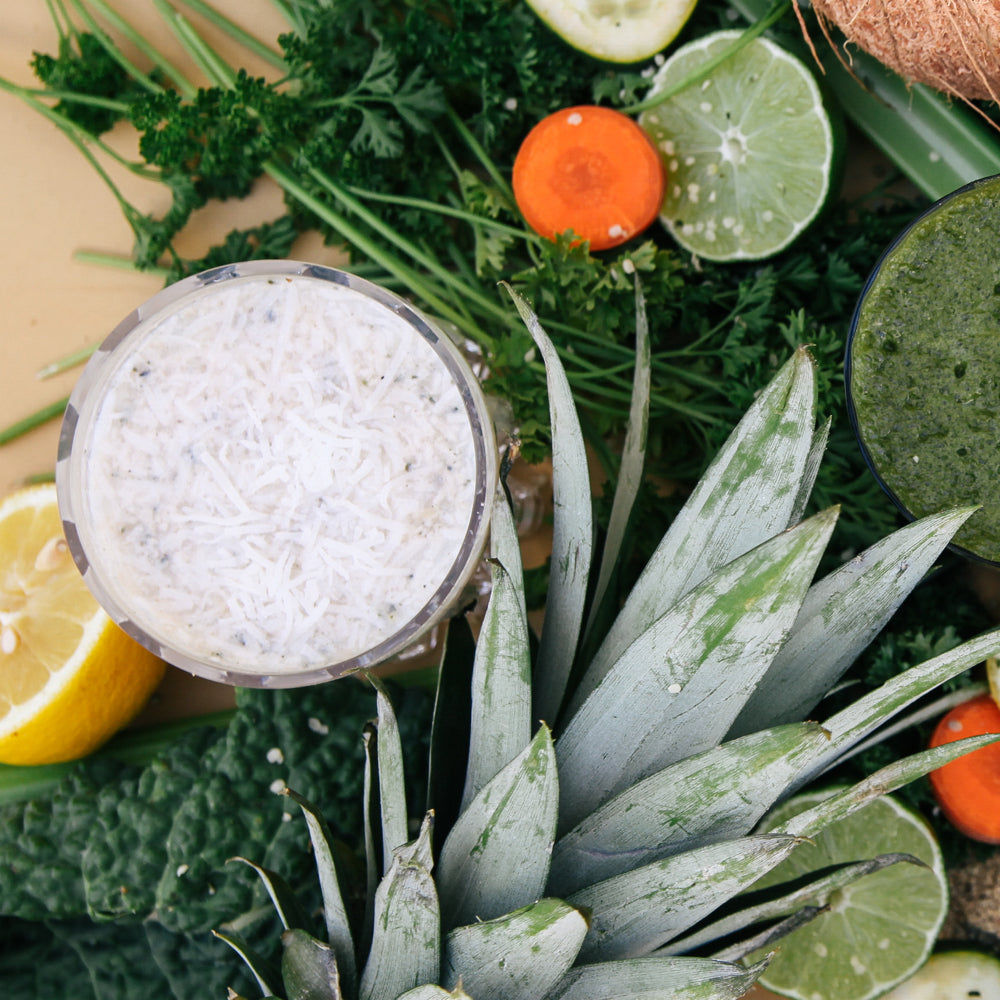 1 glass of white smoothie surrounded by variety of vegetables like parsley, slices of carrots, slices of lime and lemon, and pineapple on a wooden surface.