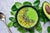 a bowl of green jalapeno pineapple smoothie sprinkled with chia seeds and nuts placed on top of a bunch of basil leaves with cut avocado and a spoon on the side