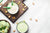 two bowls of Pistachio Smoothie, one was placed on top of a wood chopping board and a bowl of pistachio ice cream on the side of another bowl of pistachio smoothie with scattered pistachio on top of the table