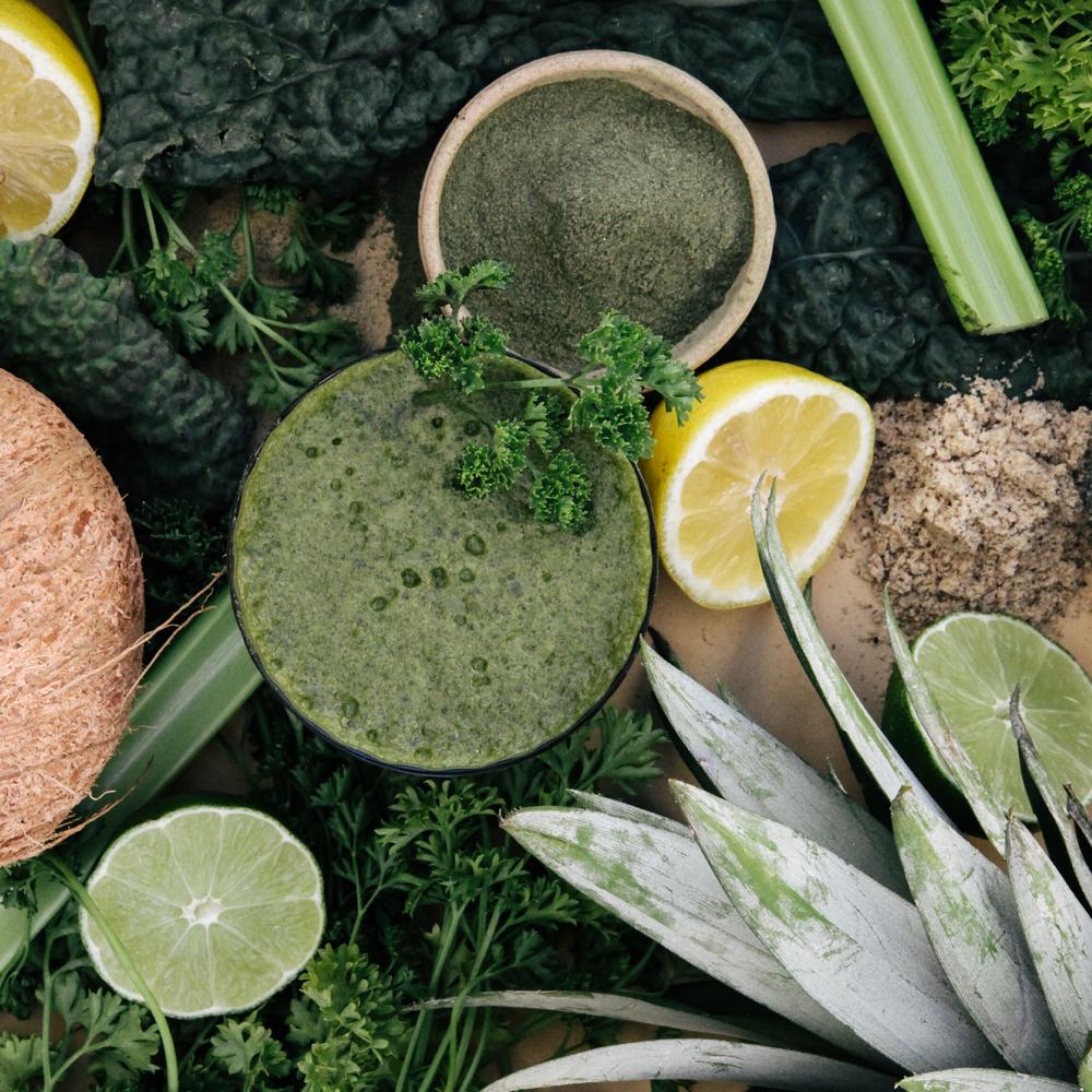 Bird-eye shot of a glass of green smoothie with a parsley garnish beside an open jar with green powder surrounded by cut lemons, cut limes, green vegetables, pineapple, and celery.